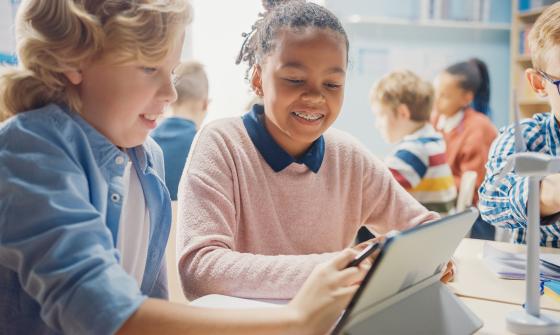 Two students in a full classroom lean over a tablet computer, smiling.