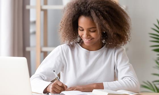 Teenager writing in a notebook next to a laptop