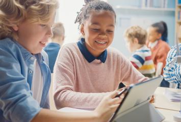 Two students in a full classroom lean over a tablet computer, smiling.