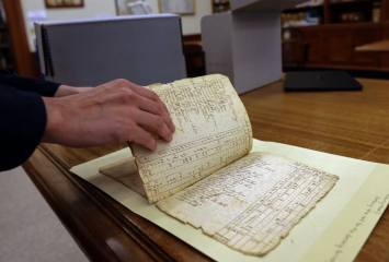A researcher's hand flips the page of a frayed and fragile, historic document.