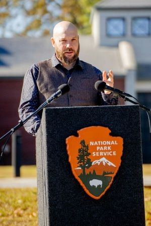 Green and Blue author Daniel Shiels Green and Blue author Daniel Shiels standing at a National Park Service podium outdoors, speaking into two microphones at either side of it.
