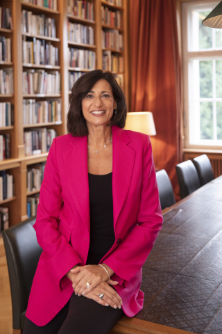 Dr. Walensky sitting on the corner of a long table with bookshelves behind her. She is smiling wearing a pink jacket with a black shirt and pants. She has brown short hair.