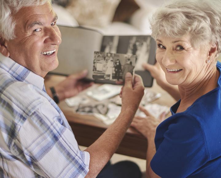 Man and woman sit at a table with array of family photos and books, looking over their shoulders and smiling