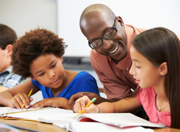 A teacher, smiling, leans down between two students at a table to see their work.