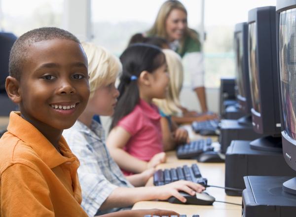 Student at computer smiles at camera; behind him, a row of students works at computers, and a teacher leans down to help at the end of the row