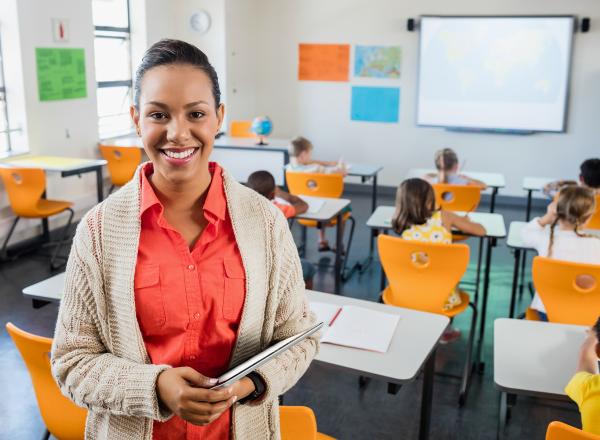 Teacher facing camera and smiling, in front of students at desks who are facing away towards the front of the classroom