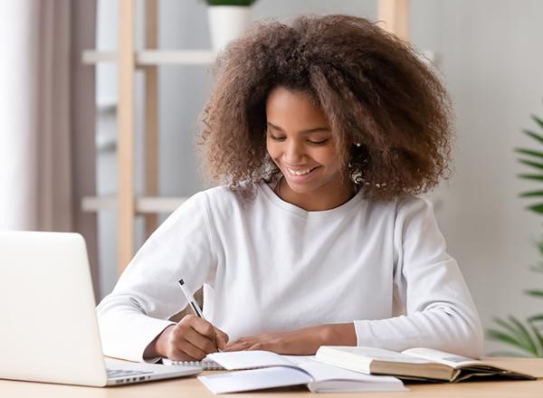 Teenager writing in a notebook next to a laptop