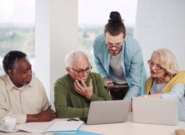 Three adult students, seated with laptops, look at screen while standing teacher leans down to point to something