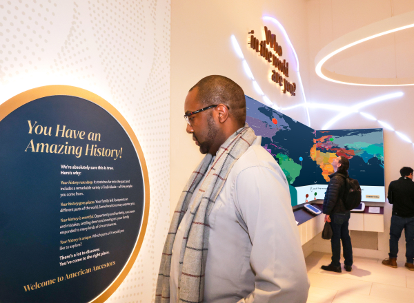 Adult male looks at a sign that reads "You have an amazing history!" in the lobby of Family Heritage Experience. In the background are three males exploring a world map. 