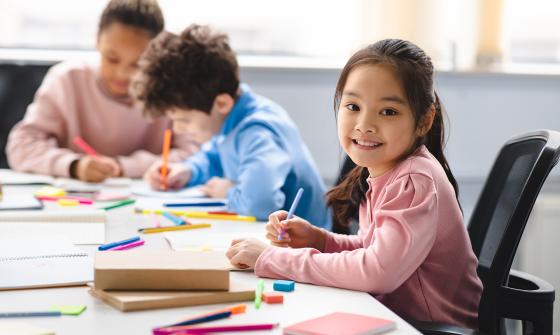 Student smiles at camera, looking up from her drawing, at table surrounded by colorful markers. Two other students focus on their drawings in the background.
