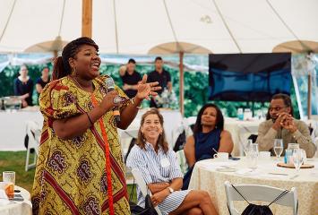 Woman standing speaks into microphone while smiling onlookers sit nearby