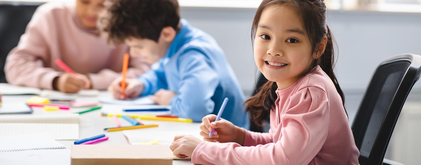 Student smiles at camera, looking up from her drawing, at table surrounded by colorful markers. Two other students focus on their drawings in the background.