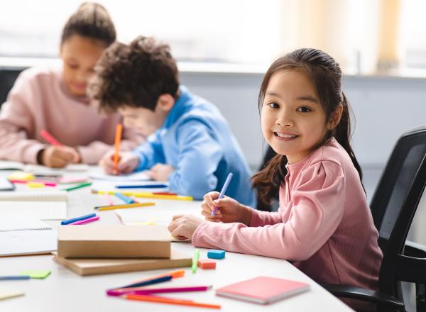Student smiles at camera, looking up from her drawing, at table surrounded by colorful markers. Two other students focus on their drawings in the background.
